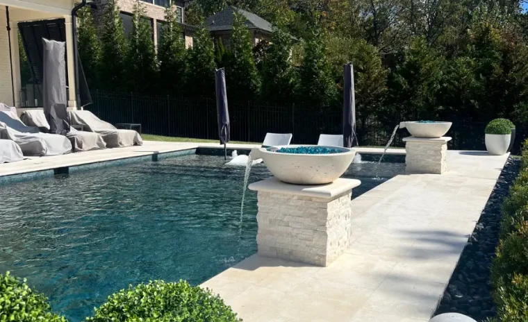 A luxurious outdoor pool area with two modern water fountains on stone pedestals. Lounge chairs with covers line one side, and neatly trimmed green bushes in large pots frame the pool.
