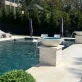 A luxurious outdoor pool area with two modern water fountains on stone pedestals. Lounge chairs with covers line one side, and neatly trimmed green bushes in large pots frame the pool.